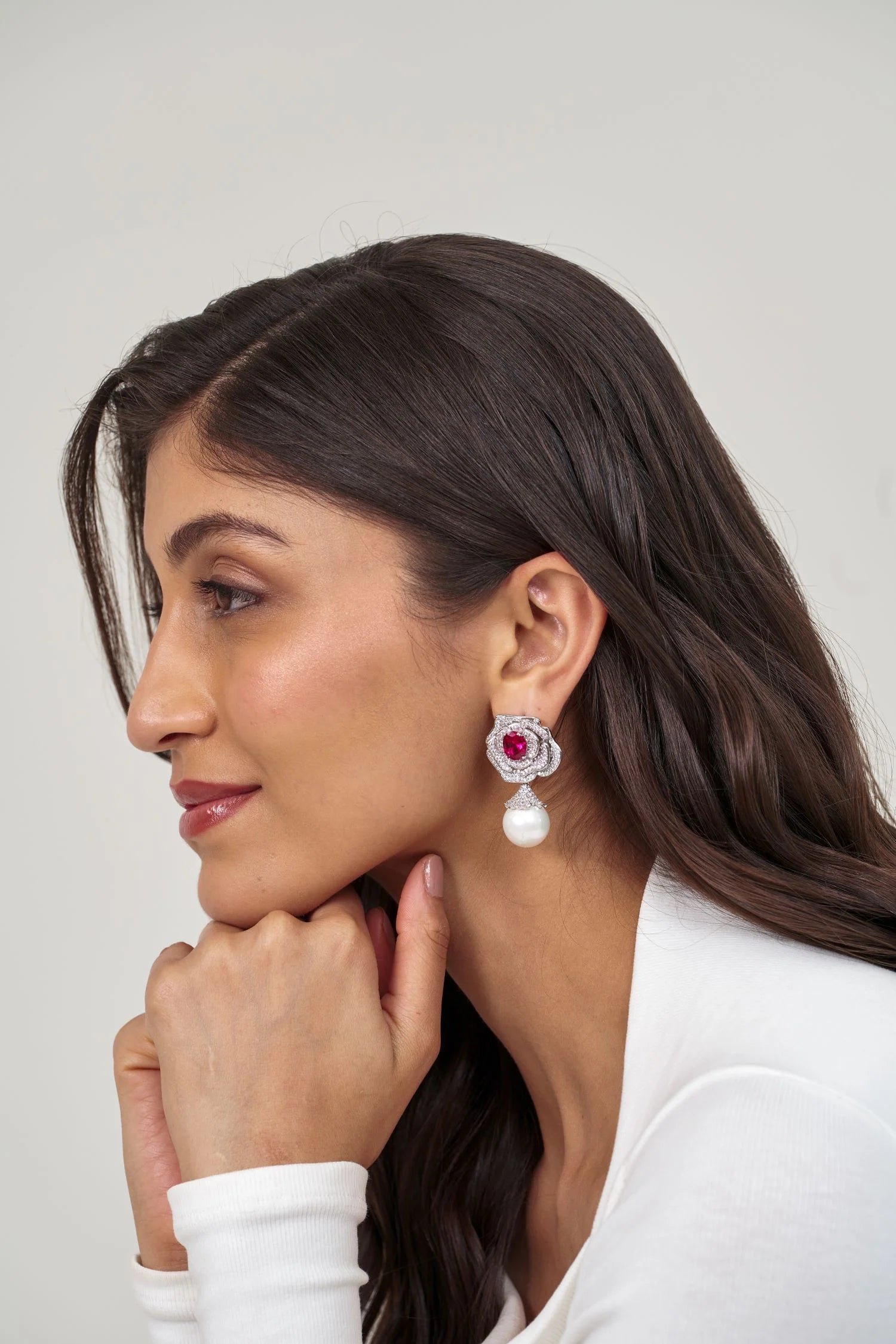 Close-up portrait of a woman wearing a floral diamond earring with a vivid pink center stone and a single pearl drop.

