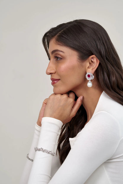 Fine jewelry close-up featuring a layered diamond floral stud with a bold pink centerpiece and suspended pearl detail.
