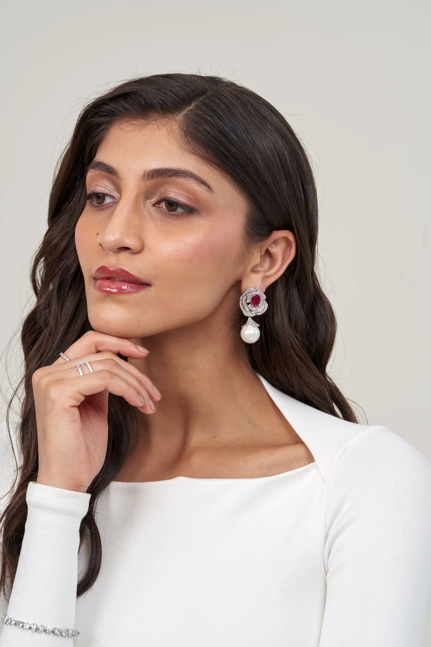 Detailed portrait of a woman wearing a statement floral earring combining a vibrant pink stone, sparkling halo, and pearl pendant.
