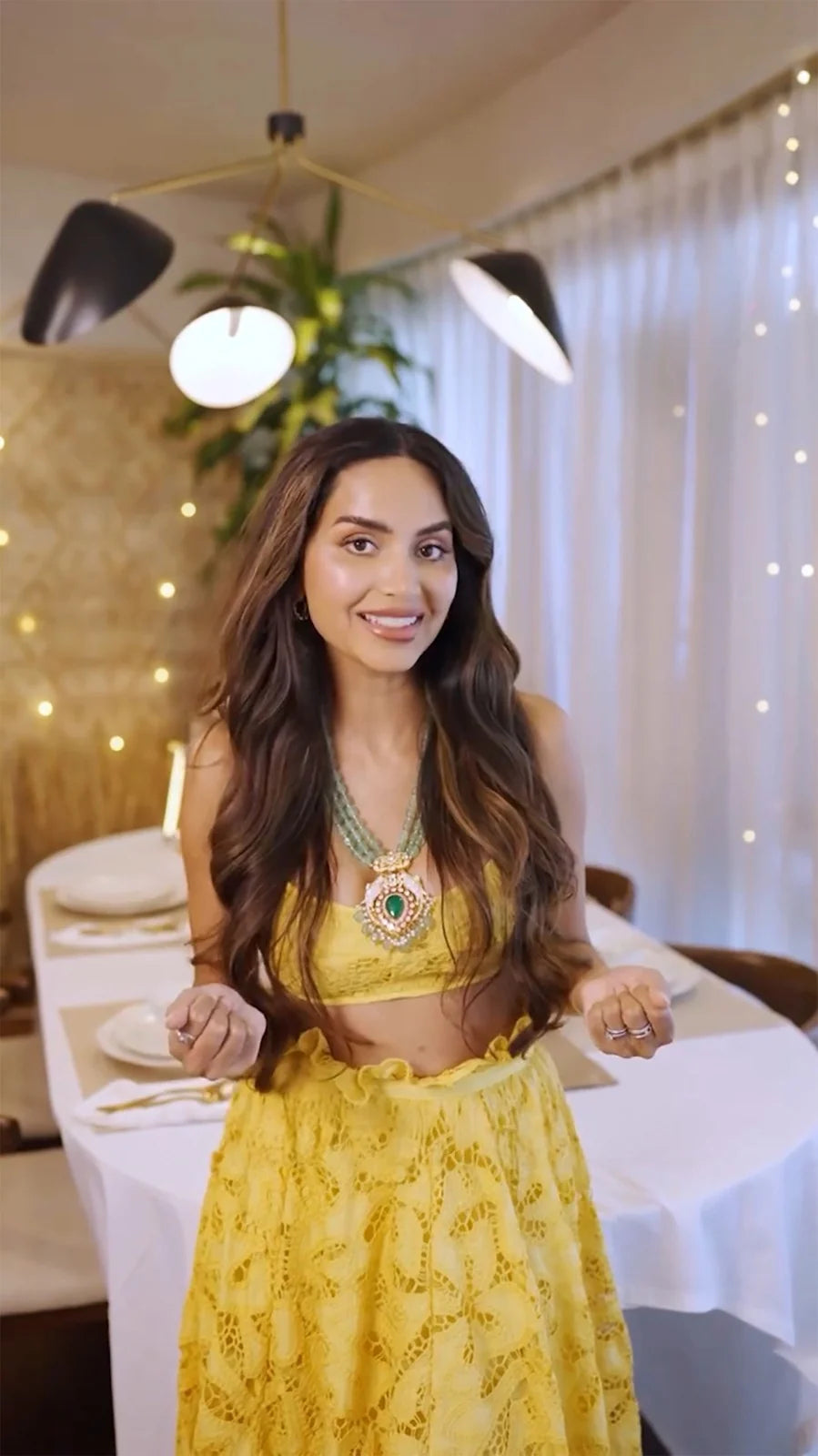 Close-up portrait of a woman in a yellow outfit wearing a layered green gemstone necklace with a bold statement pendant in a warm indoor setting.
