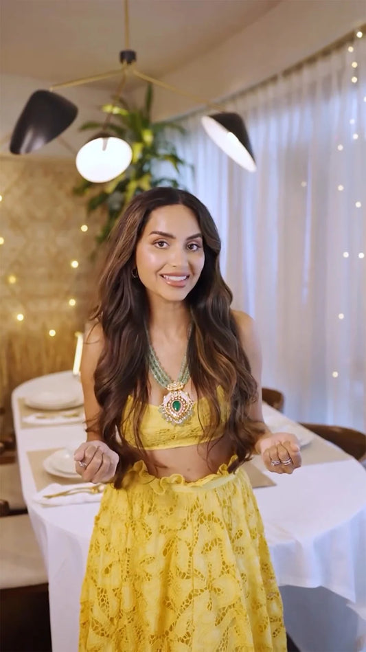 Close-up portrait of a woman in a yellow outfit wearing a layered green gemstone necklace with a bold statement pendant in a warm indoor setting.

