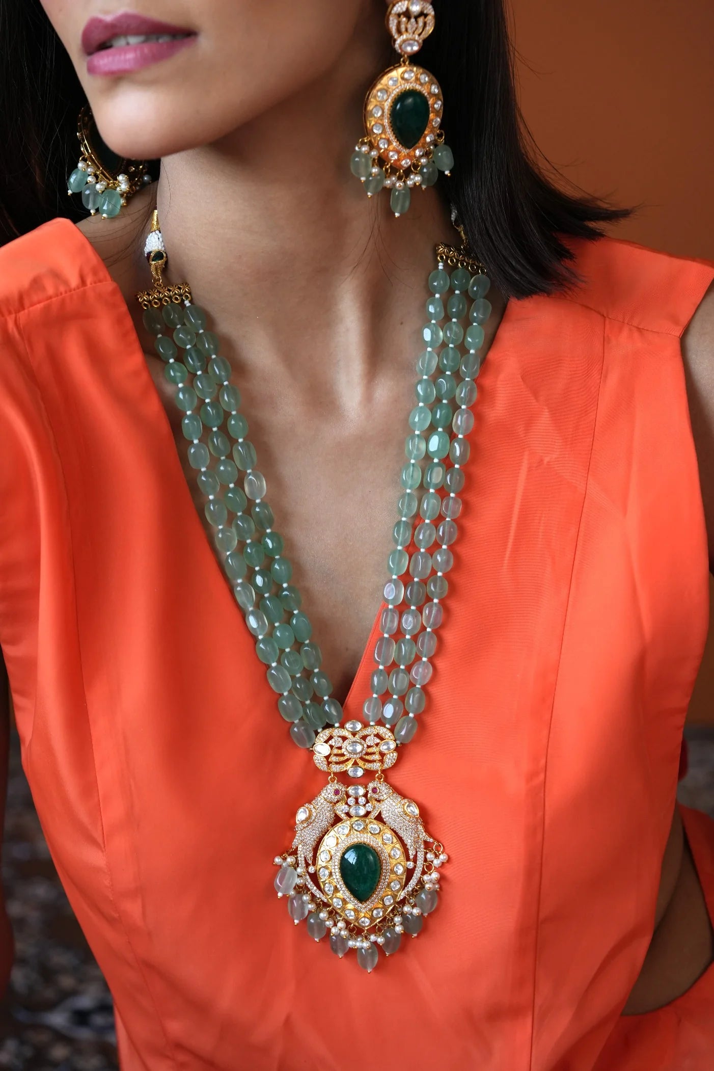 Elegant lifestyle shot of a smiling woman styled in a yellow lace outfit with a multi-layer necklace featuring a large green centerpiece.
