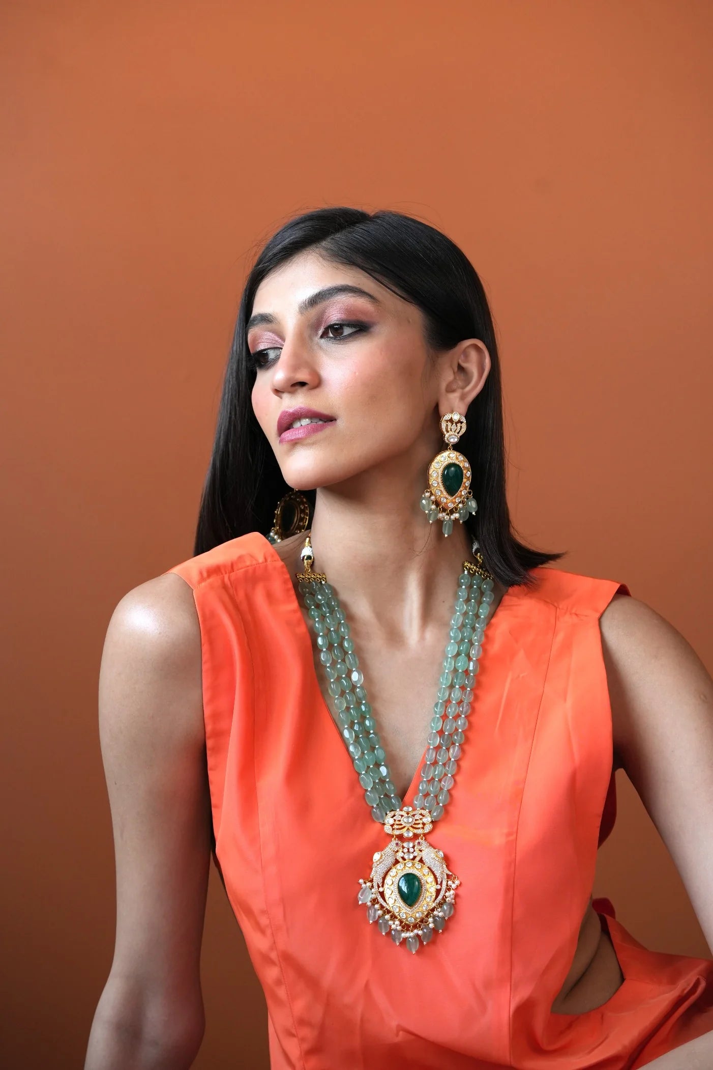 Indoor styling image of a woman adorned with a statement gemstone necklace and matching layers, set against a softly lit background.
