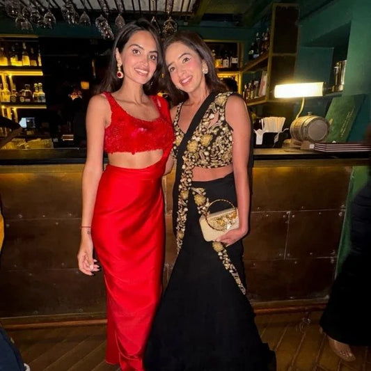Close-up of two women dressed in glamorous evening wear, styled with earrings and a handbag in a warm indoor setting.
