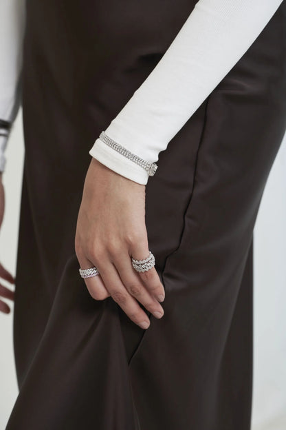 Close-up of a hand wearing stacked diamond rings and a slim diamond bracelet, styled with a white sleeve and dark fabric contrast.

