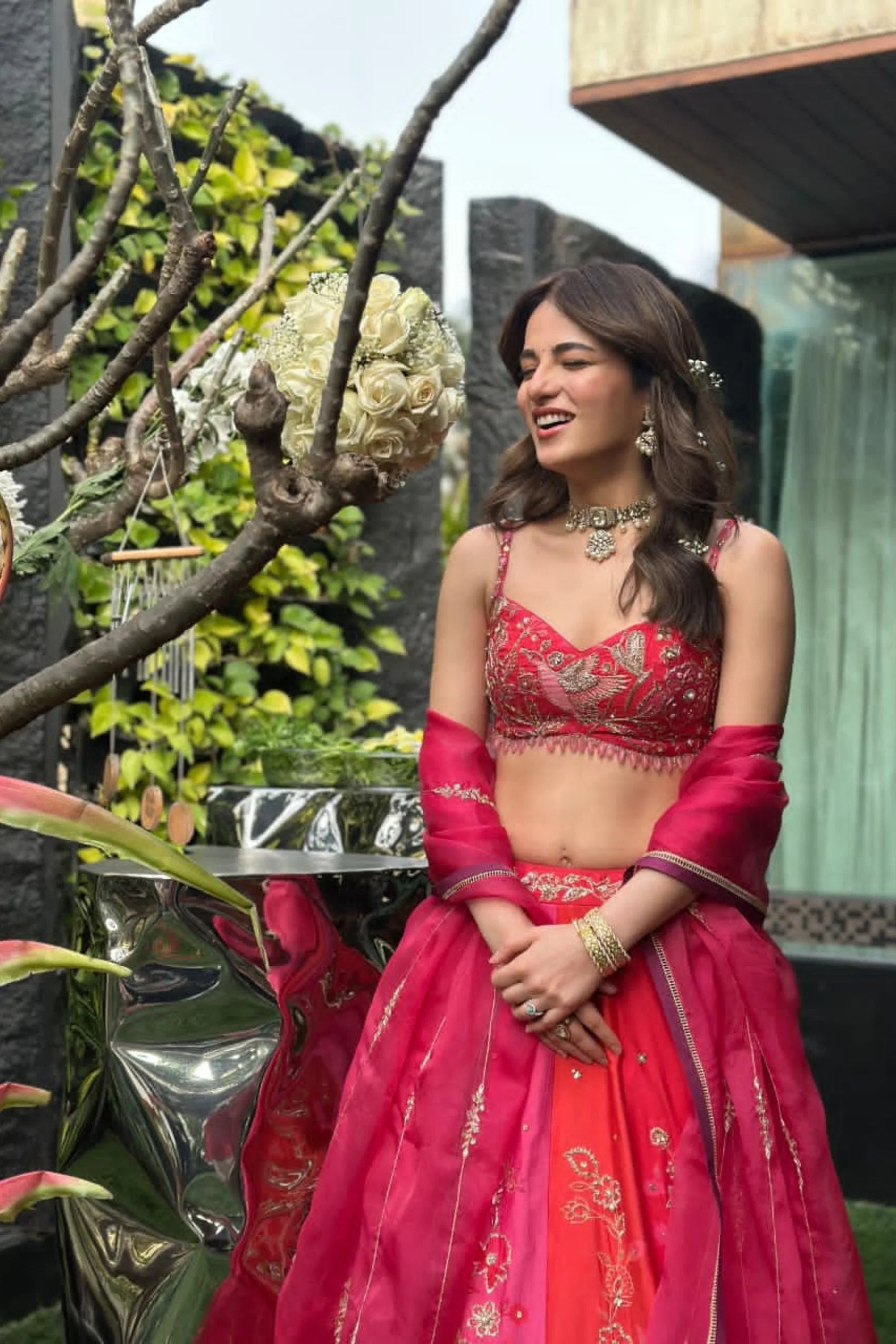Close-up of a woman in a red embroidered lehenga with a matching dupatta, wearing a statement choker necklace and ornate earrings, seated in a lush garden setting.
