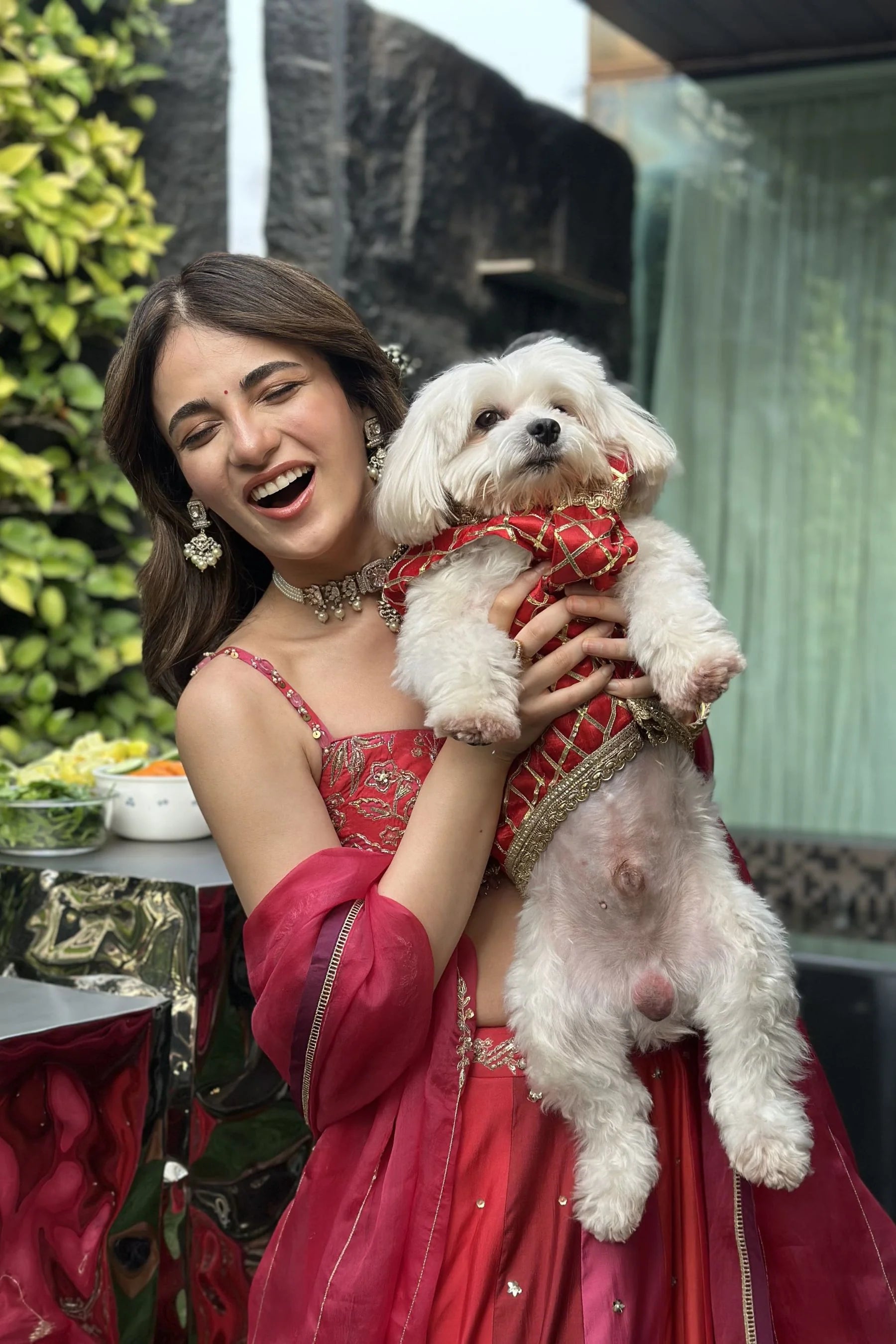 Elegant outdoor portrait of a woman in a richly detailed red outfit, styled with traditional jewelry and soft floral surroundings.
