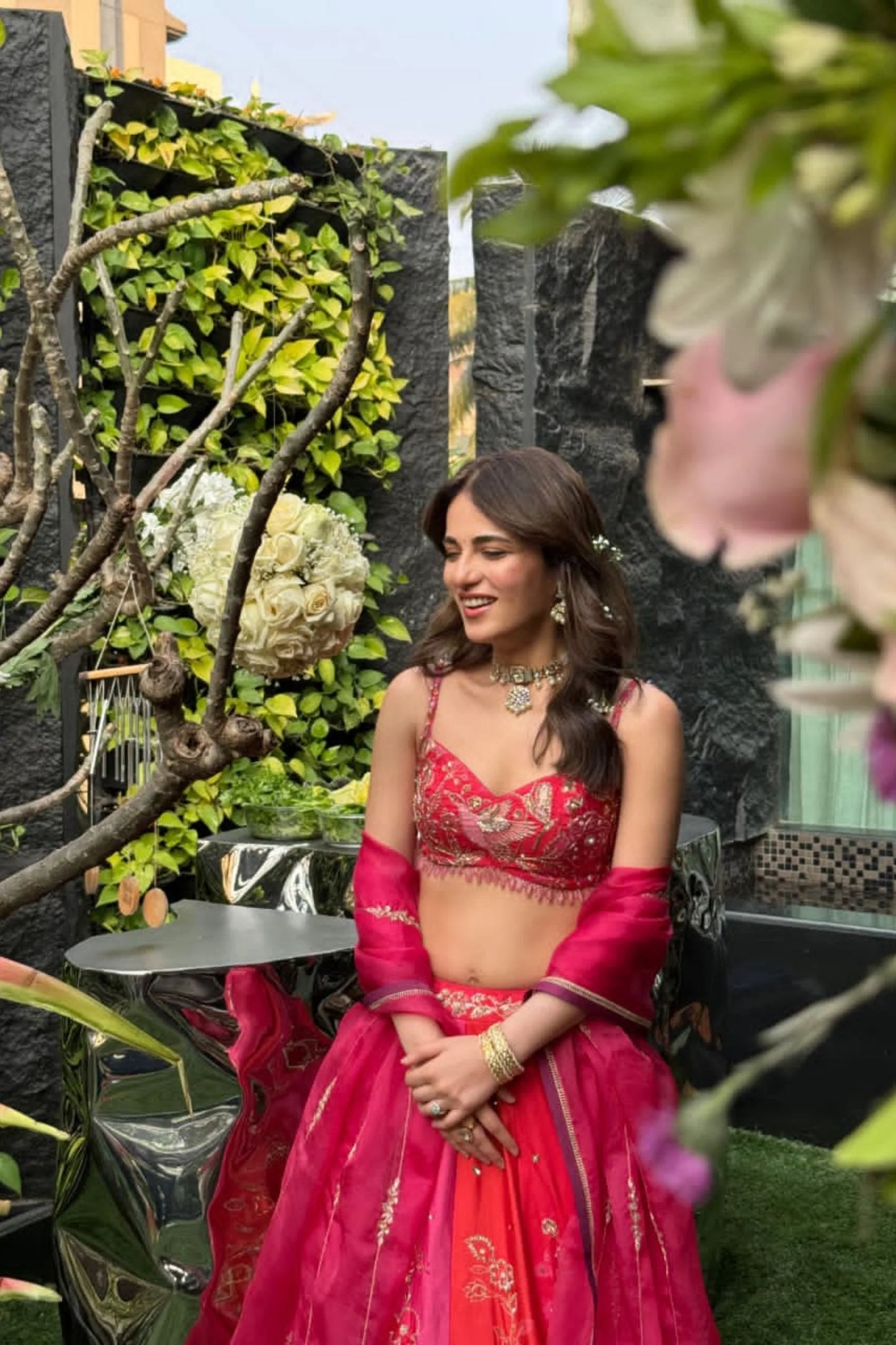 Sophisticated portrait showcasing a woman in a red outfit with delicate detailing, accessorized with a choker necklace and earrings against greenery.
