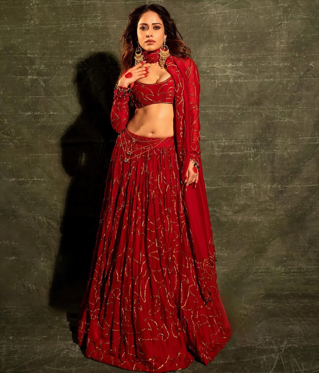 Woman in red lehenga choli with gold embroidery and statement jewelry against textured backdrop
