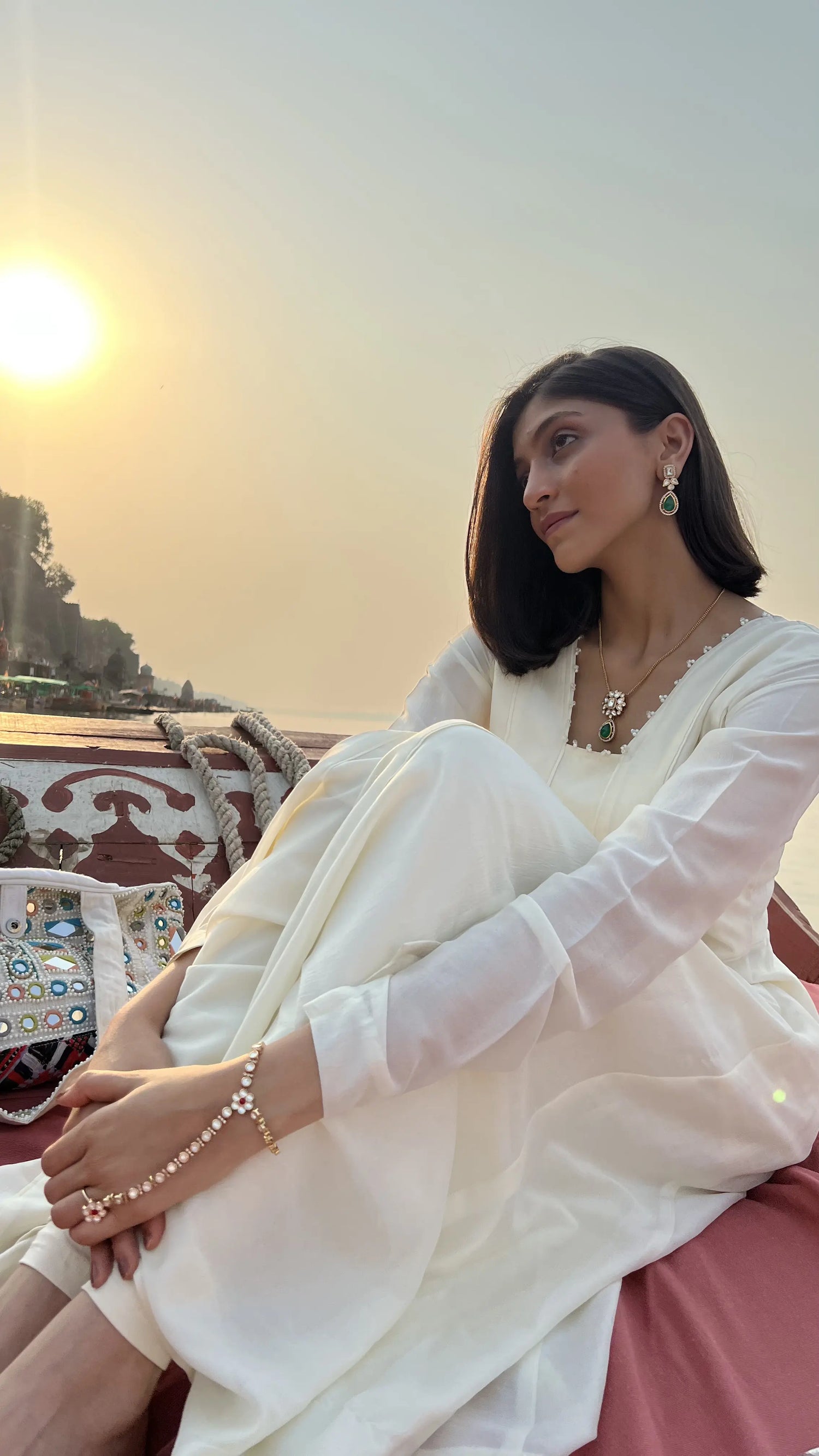 Elegant outdoor portrait of a woman in a soft white dress, styled with green stone jewelry, seated on a wooden boat at golden hour.
