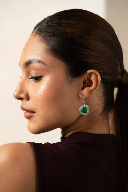 Perspective view of a heart-cut emerald earring with a surrounding diamond halo and delicate drop setting.